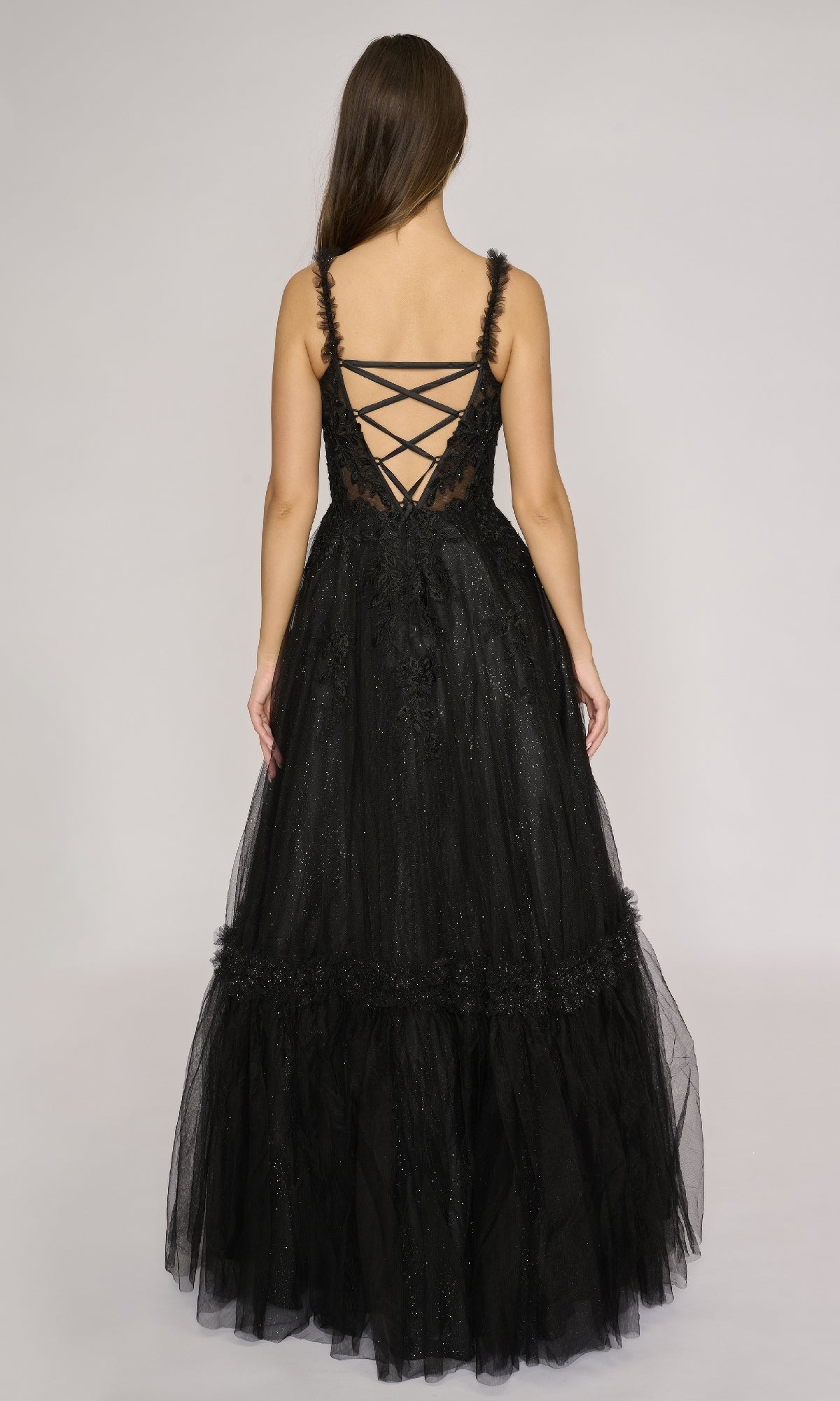 A woman models the Nina Canacci 3272 long formal dress with a sleeveless lace bodice and glitter tulle A-line skirt, standing against a plain light background and facing away from the camera.